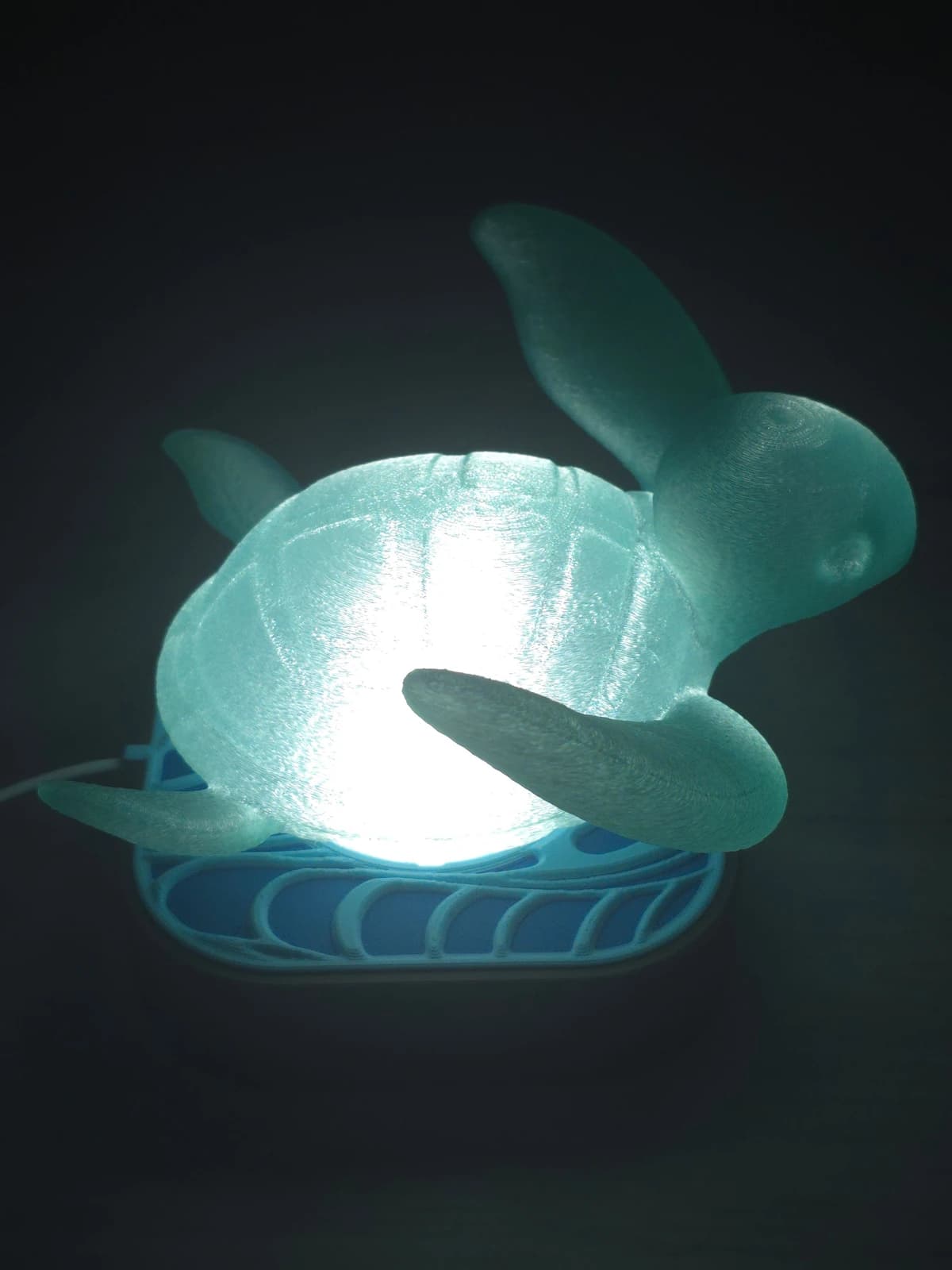 Lampa nocna z kloszem Turtle, włączona — żółw świeci nad podstawą z wzorem fal